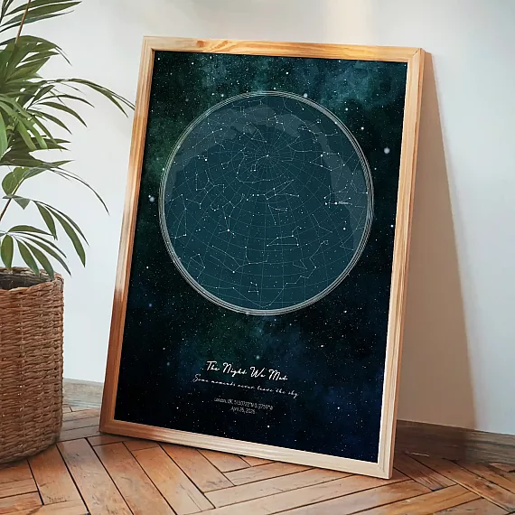 Framed personalized star map print leaning beside a plant on wooden flooring.