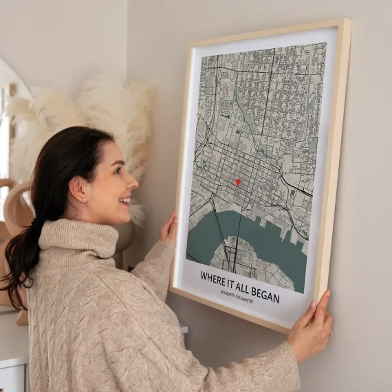 Woman hanging a Where It All Began custom map poster with a heart marking a special location on the wall.