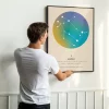 Mężczyzna wiesza oprawiony plakat zodiakalny Bliźnięta z kolorową aurą, konstelacją i astrologicznymi detalami.